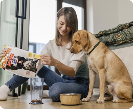 Žena nasypáva krmivo značky Hill's Pet Nutrition do odmerky, zatiaľ čo pes ju pozorne sleduje.